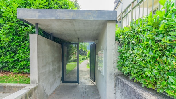 A narrow concrete pathway leading to a building entrance, surrounded by lush green bushes. - Sunflowers Home - by Varenna Holidays (Mandello del Lario)