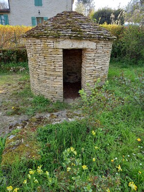 A stone structure with a small opening, surrounded by greenery and a building in the background.