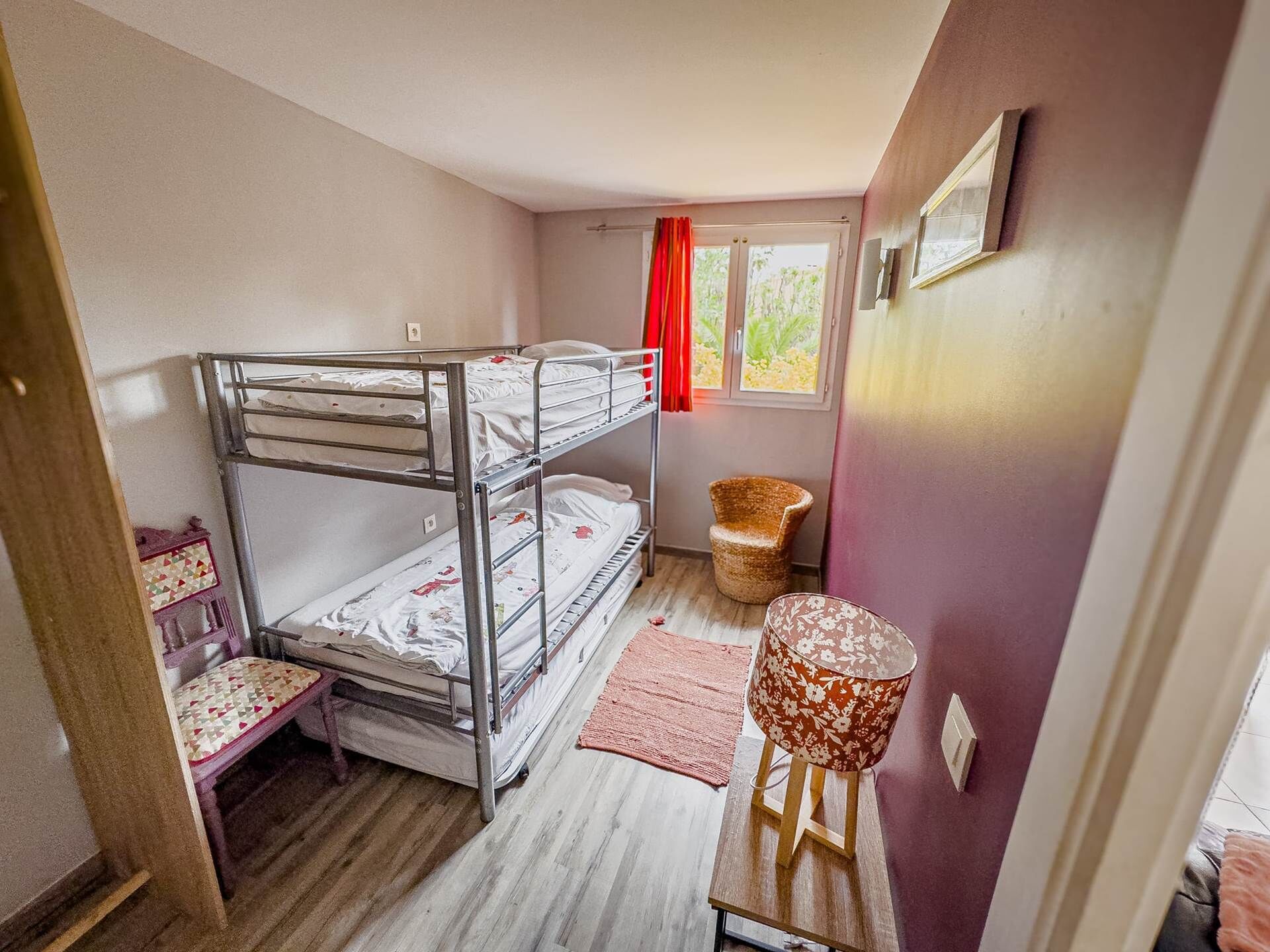 Ein Schlafsaalzimmer mit Etagenbetten, einem kleinen Tisch mit Lampe, einem Korbstuhl und einem Fenster mit roten Vorhängen.