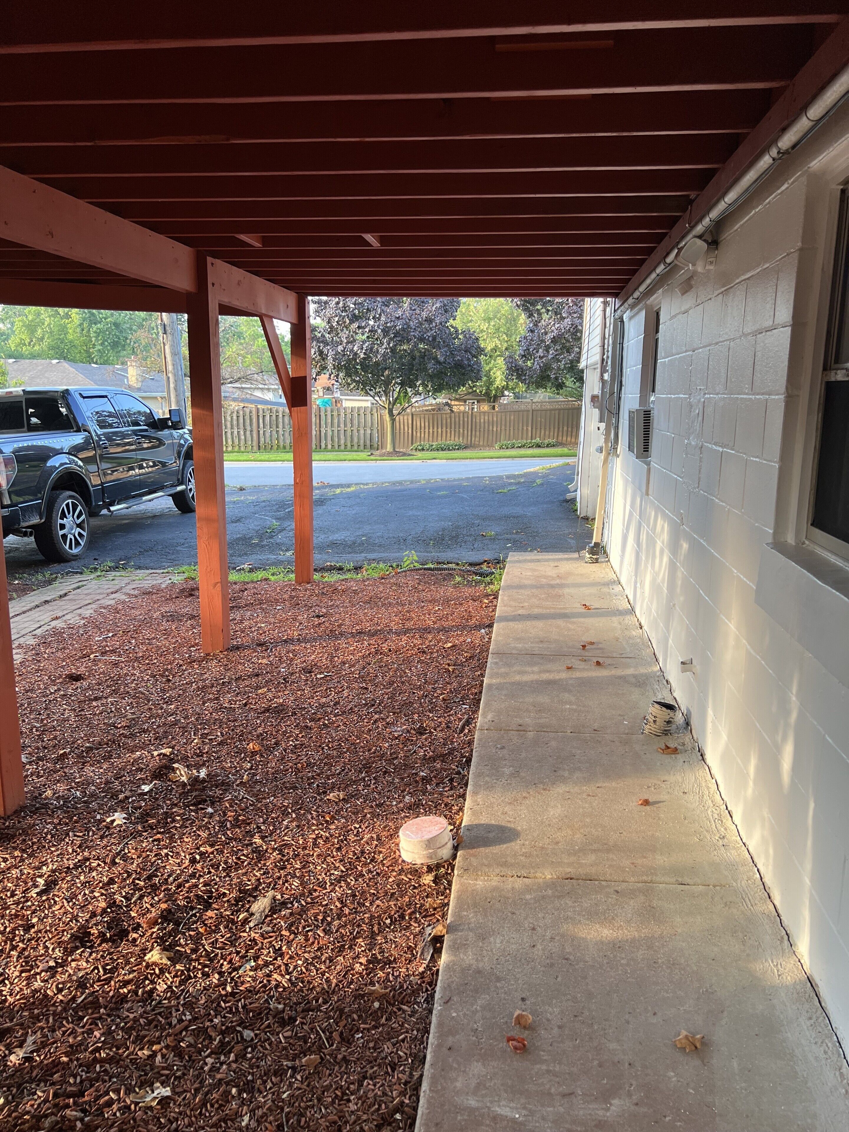 A covered patio area with a concrete walkway, wooden deck, and a vehicle parked in the background.