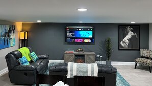 A modern living room with a black leather sofa, a flat-screen TV mounted on the wall, a fireplace, and a framed artwork.