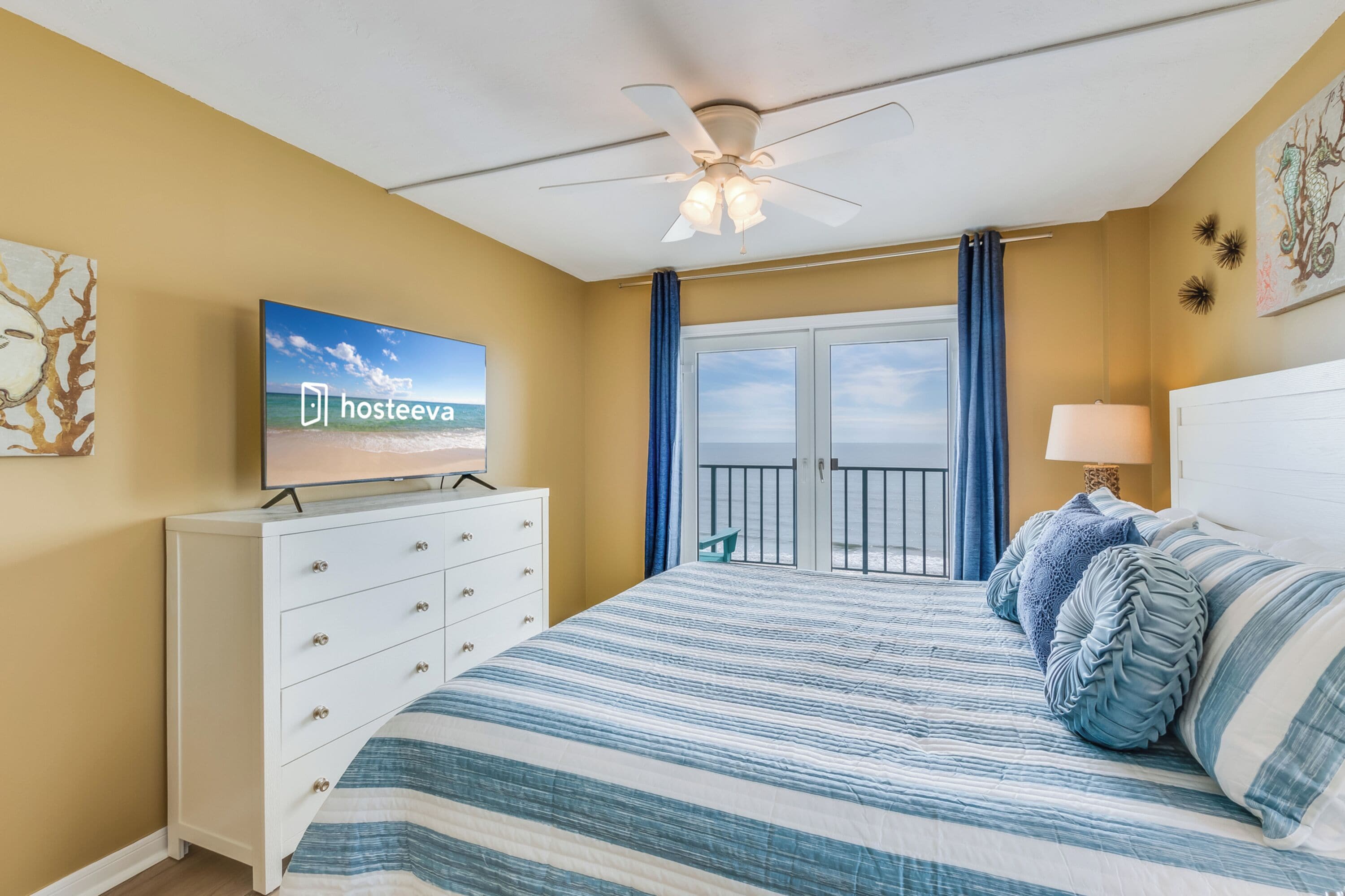 A bedroom with a bed, a dresser, a ceiling fan, a TV, and a balcony with curtains.