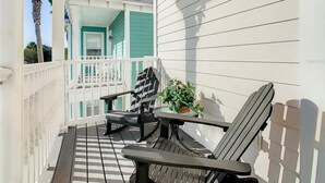 A covered porch with two chairs and a small table, a white railing, and a view of a building with teal siding. - Reunion Resort 3 bed With Private Pool (Kissimmee)