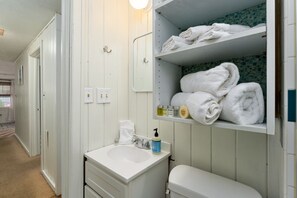 A bathroom with a white sink, a mirror, a towel rack, and a shelf with folded towels. - Unique Getaway Steps to Wrightsville Beach! (Wrightsville Beach)
