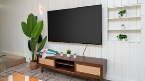 Una sala de estar con un televisor de pantalla plana grande montado en una pared blanca, un mueble de madera para entretenimiento con pequeñas macetas y una planta verde grande en una cesta tejida.