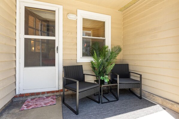 A porch with two black wicker chairs, a potted plant, and a welcome mat.