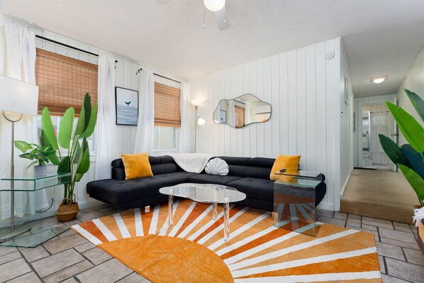 A modern living room with a black sofa, glass coffee table, and orange patterned rug. - Unique Getaway Steps to Wrightsville Beach! (Wrightsville Beach)