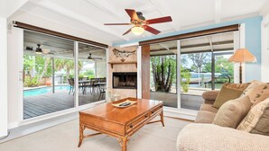 Un salon spacieux avec une table basse en bois, un canapé et un ventilateur de plafond. De grandes fenêtres offrent une vue sur une terrasse extérieure avec une piscine et du mobilier.