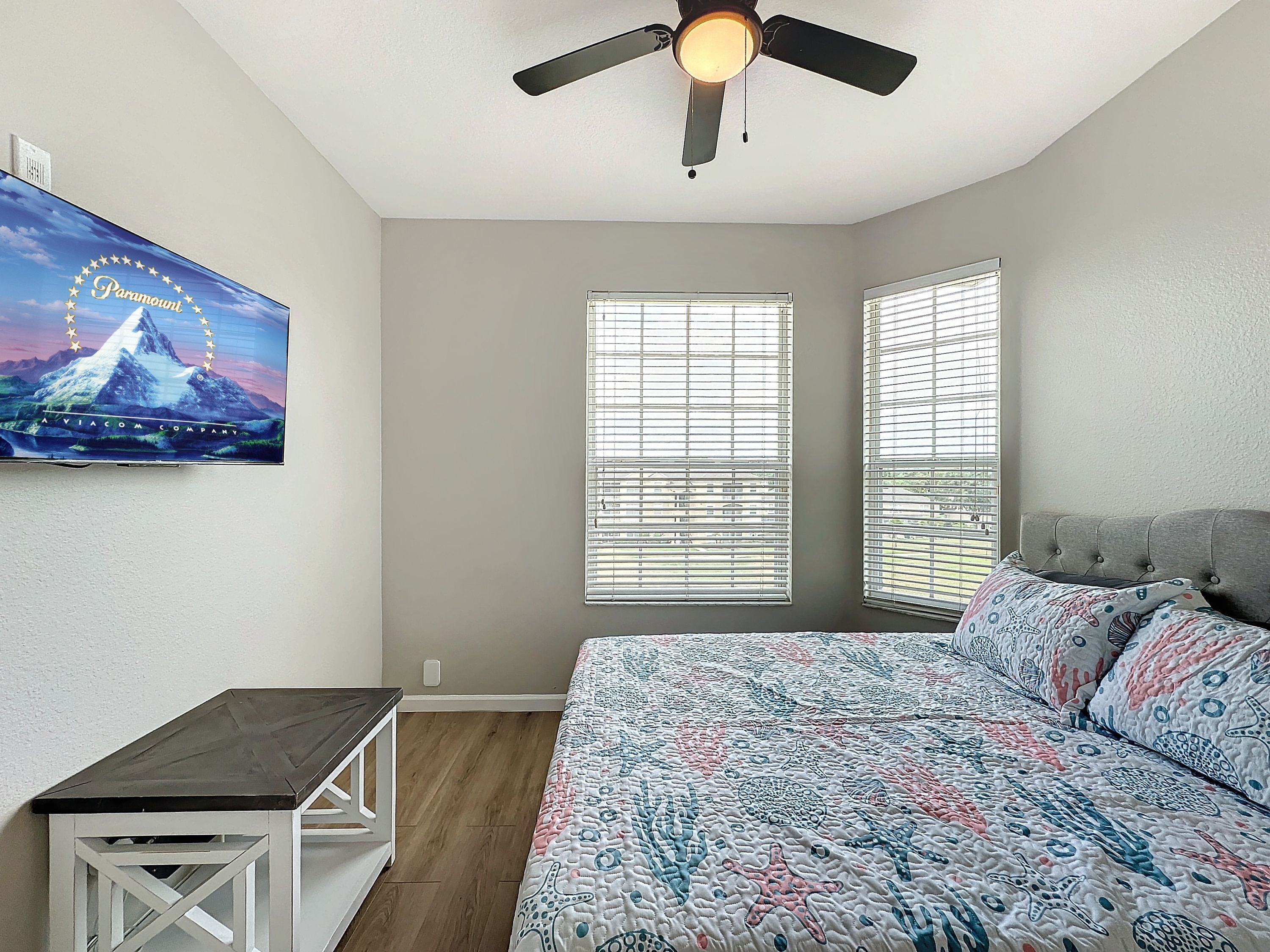 A bedroom with a bed, a ceiling fan, two windows with blinds, a small table, and a wall-mounted TV displaying a Paramount movie poster.