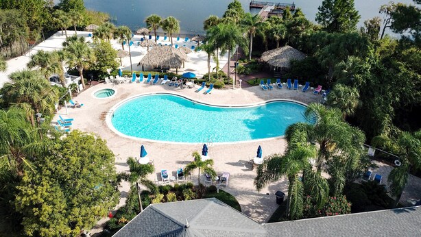 Een resort met een groot zwembad, omringd door ligbedden en parasols, met op de achtergrond een meer en een dichte boomgrens.