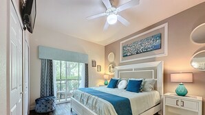 Een slaapkamer met een bed, een plafondventilator, een raam met gordijnen, een nachtkastje en een bedtafeltje.