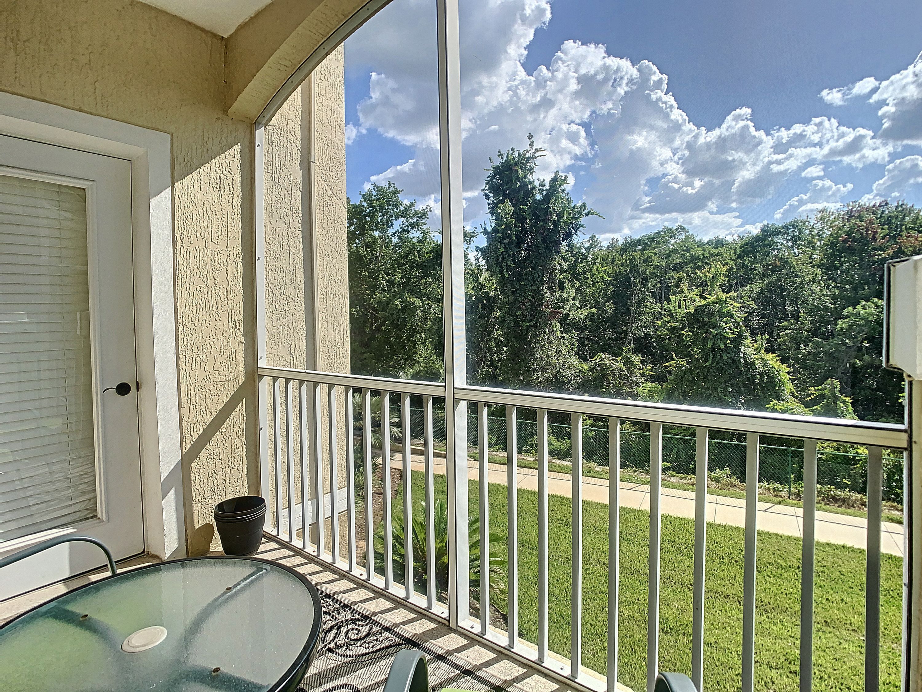 Un balcon avec une table et des chaises en verre, une porte et une vue sur une pelouse verdoyante et des arbres.