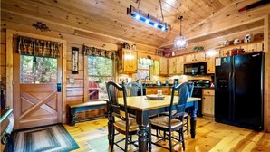A wooden cabin interior with a dining table, chairs, a kitchen area, and a large window. - Cozy Cabin near DT Blue Ridge, GA 🌲Mtn Views💧Hot Tub 🔥Fire Pit 🐶P... (Blue Ridge)