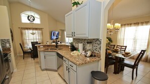 Una cocina con encimeras de granito, armarios blancos, un comedor con mesa y sillas, y un televisor montado en la pared.