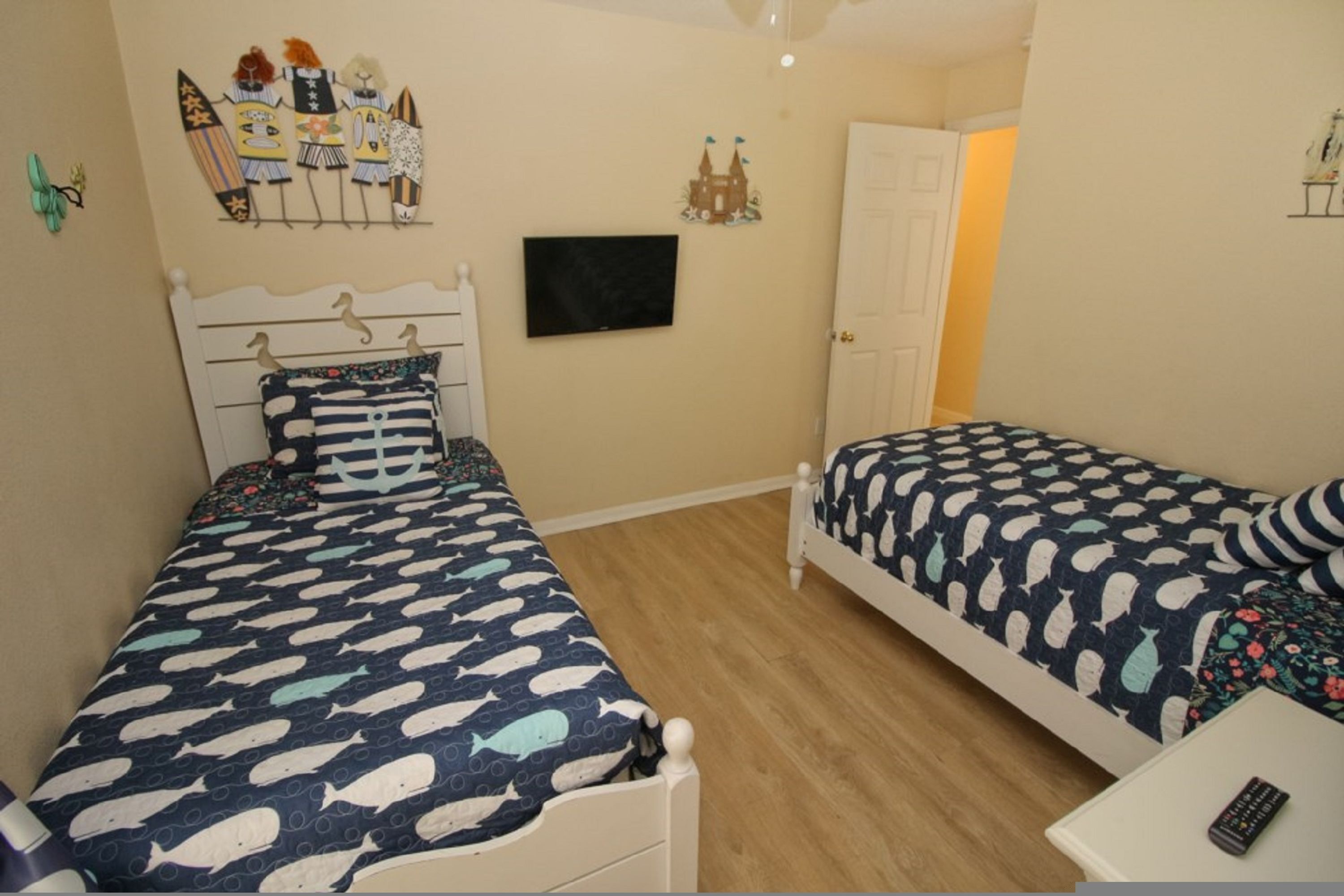 Une chambre à coucher avec deux lits, chacun recouvert d’une couvre-lit à motifs de poissons bleus et blancs. Un téléviseur est fixé au mur, il y a une porte blanche et une petite table avec une télécommande.