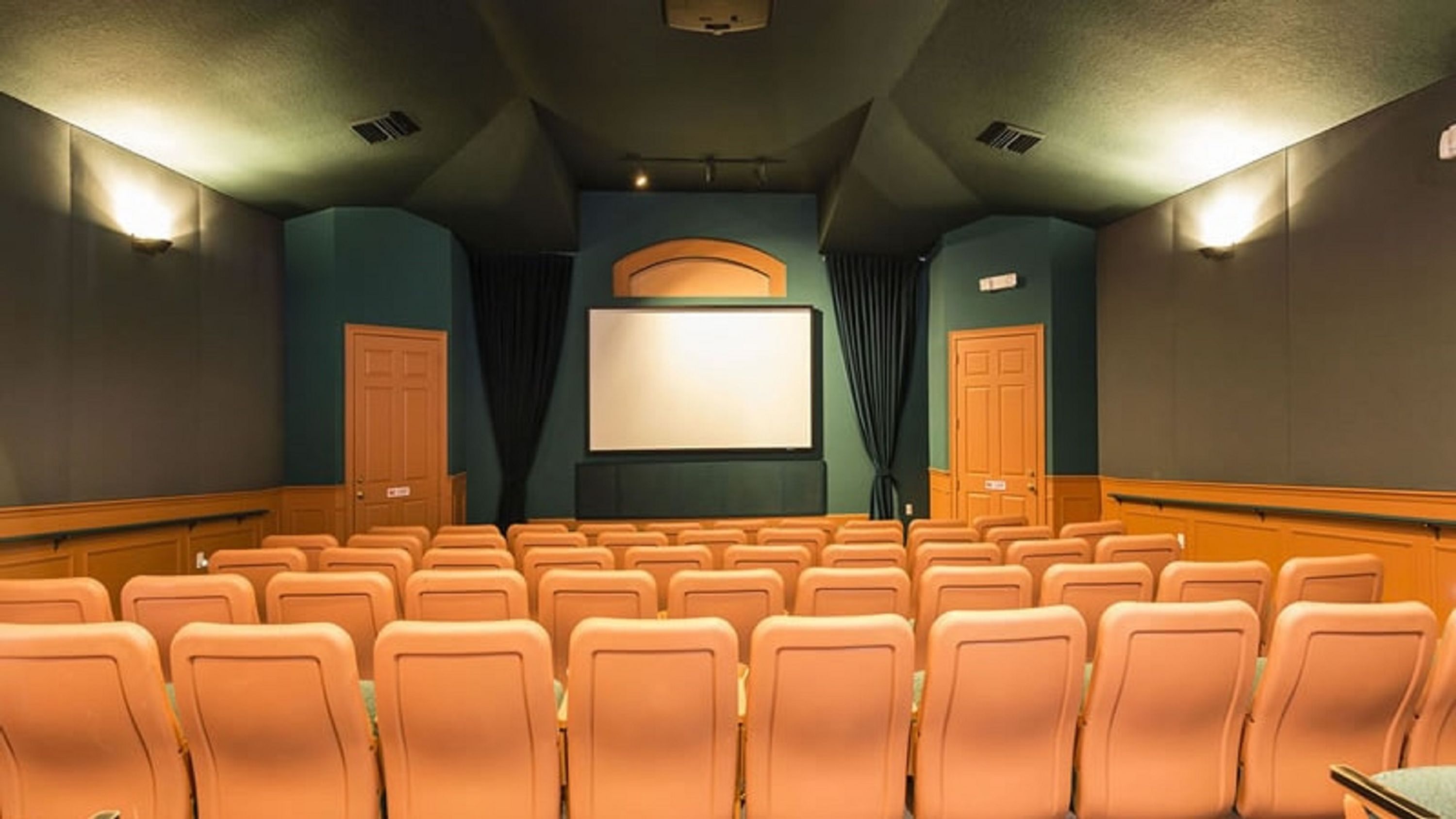 Un teatro con hileras de asientos anaranjados, una pantalla grande y dos puertas a cada lado.