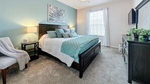 Ein Schlafzimmer mit einem Bett, einem Nachttisch, einem Sessel, einem Deckenventilator, einem Fenster mit Vorhängen und einer Kommode.