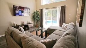 A living room with a large sectional sofa, a flat-screen TV mounted on the wall, a potted plant, and a sliding glass door leading to a pool. - Newly Renovated Home With Calming Atmosphere (Kissimmee)