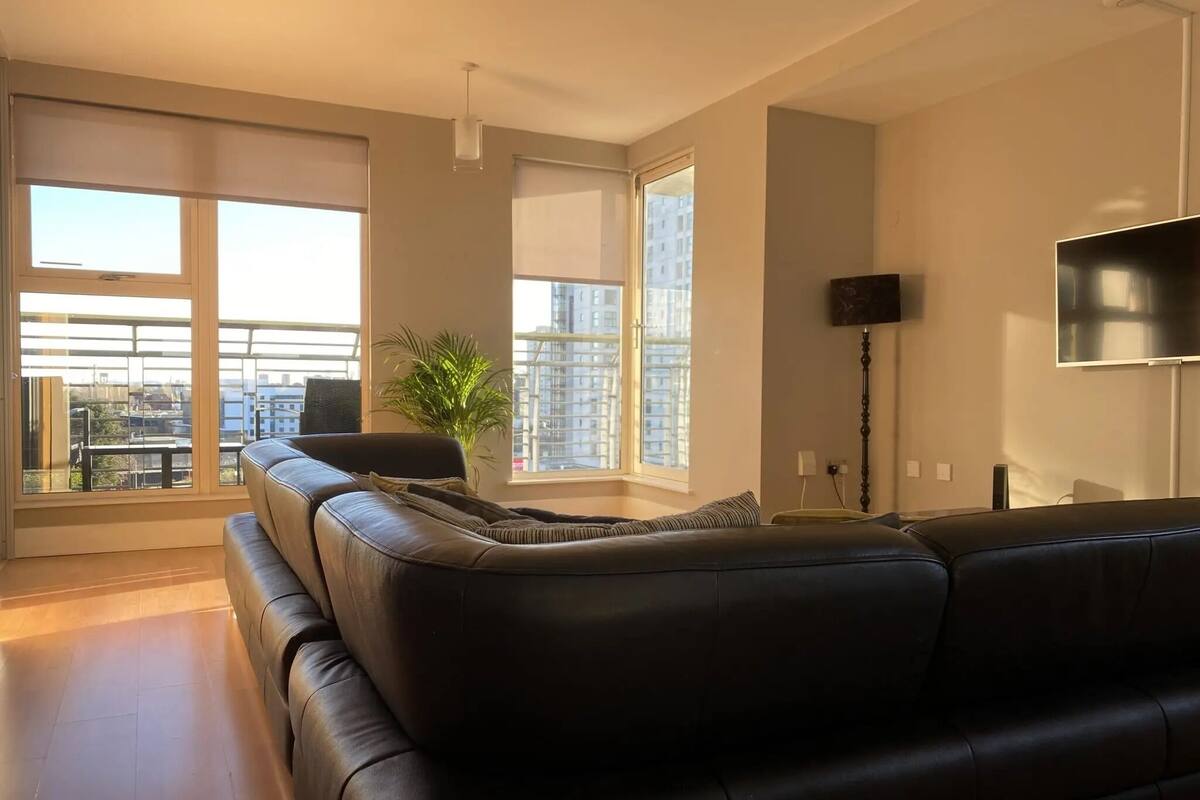 Un salon moderne avec un canapé en cuir noir, de grandes fenêtres donnant sur la ville, un lampadaire et un téléviseur à écran plat fixé au mur.