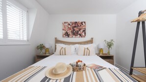 Une chambre à coucher bien rangée, avec un lit, une table de chevet dotée d’une lampe, une œuvre d’art murale et une fenêtre avec des stores.
