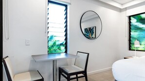 Une pièce comprenant un lit, une table, deux chaises, une fenêtre avec des stores, un miroir fixé au mur et une petite étagère.