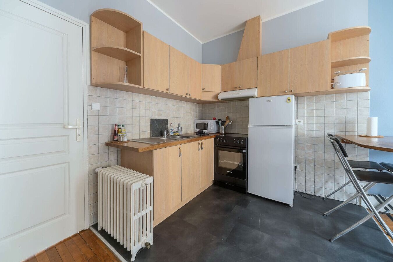 Een compacte keuken met houten kastjes, een ingebouwde oven, een koelkast en een klein eethoekje met een stoel.