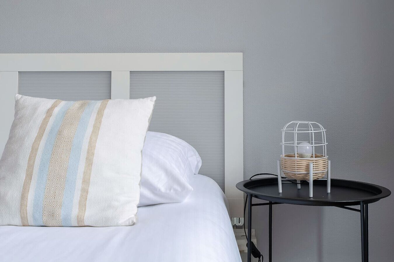 A bed with white bedding and striped pillows, a small round table with a wicker basket and a wire cage, and a grey wall.