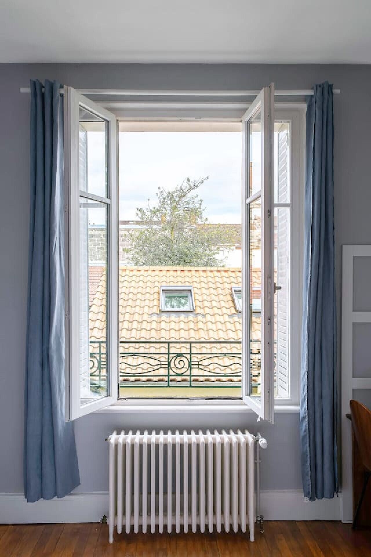 Ein Zimmer mit großem Fenster, blauen Vorhängen, einem Heizkörper und Blick auf Gebäude und Bäume.