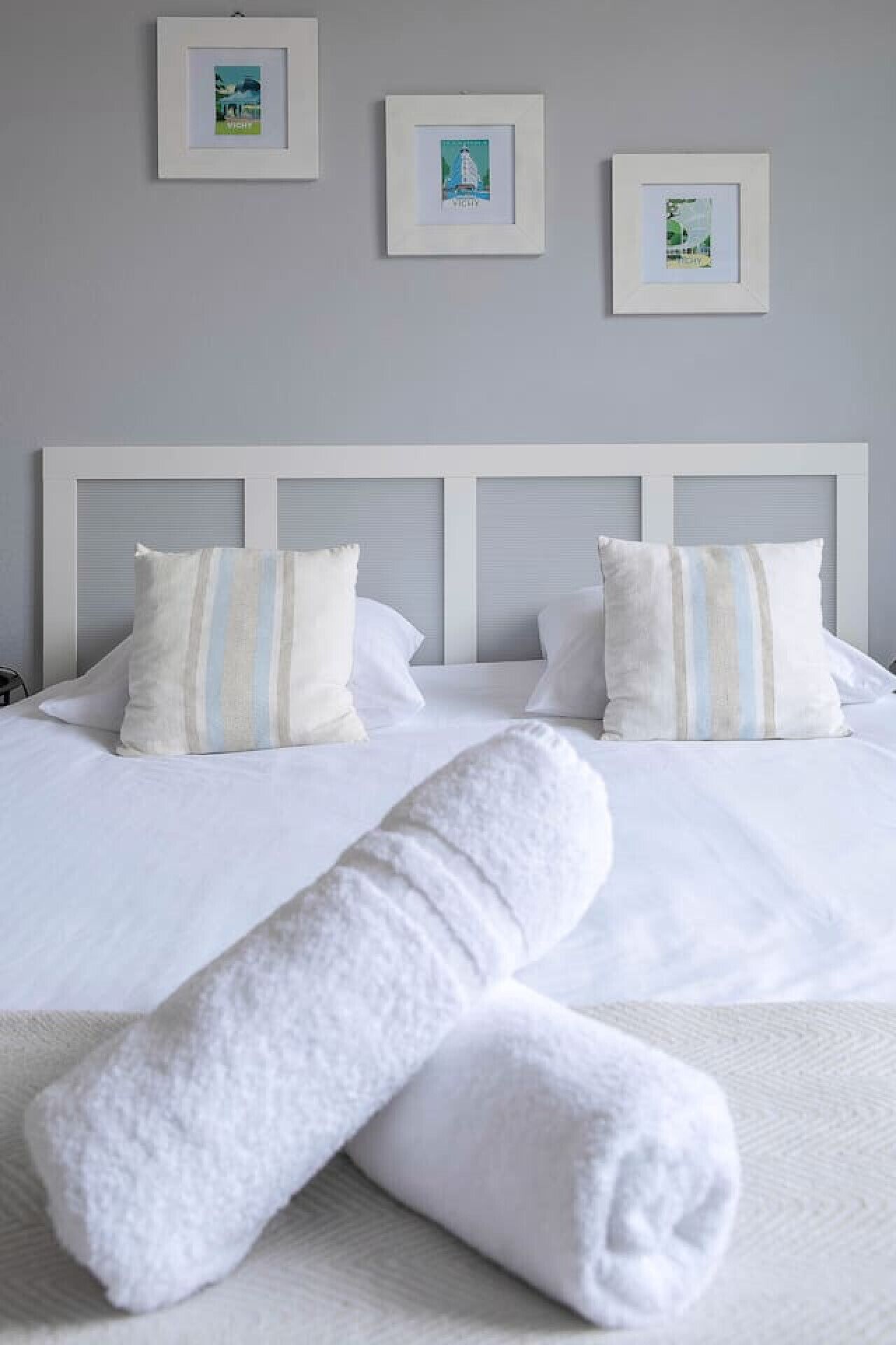 A neatly made bed with white linens and striped pillows, a rolled white towel, and three framed pictures on the wall above.