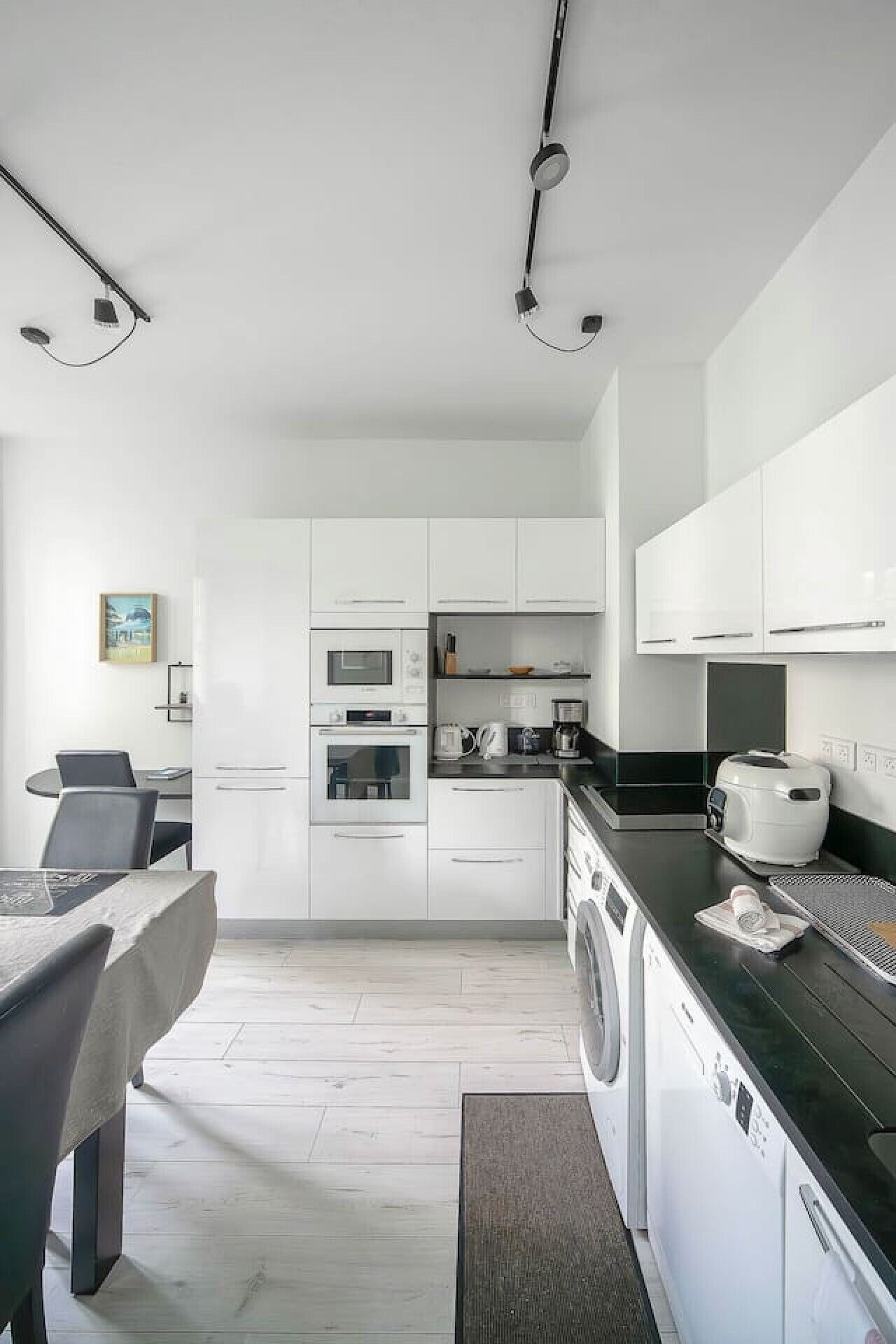Une cuisine moderne avec des meubles blancs, un plan de travail noir, un four à micro-ondes, un four traditionnel, une machine à café et un lave-linge.