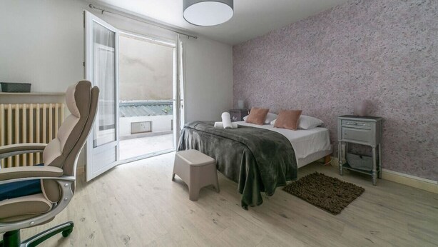 A bedroom with a bed, a chair, a stool, a nightstand, and a view of the outside.