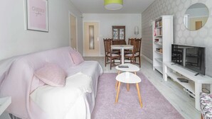 A modern living room with a sofa, a dining table with chairs, a small white table, a bookshelf, a TV, and a mirror.