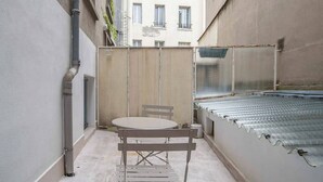 A narrow outdoor space with a table and chairs, a metal grill, and a building in the background.