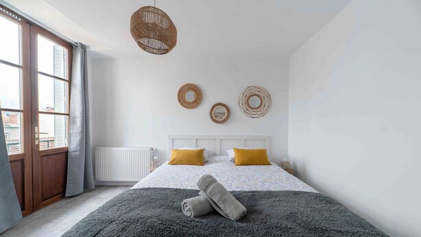 Un dormitorio con un ventanal grande, una cama con una manta gris, dos almohadas amarillas y un cabecero blanco.