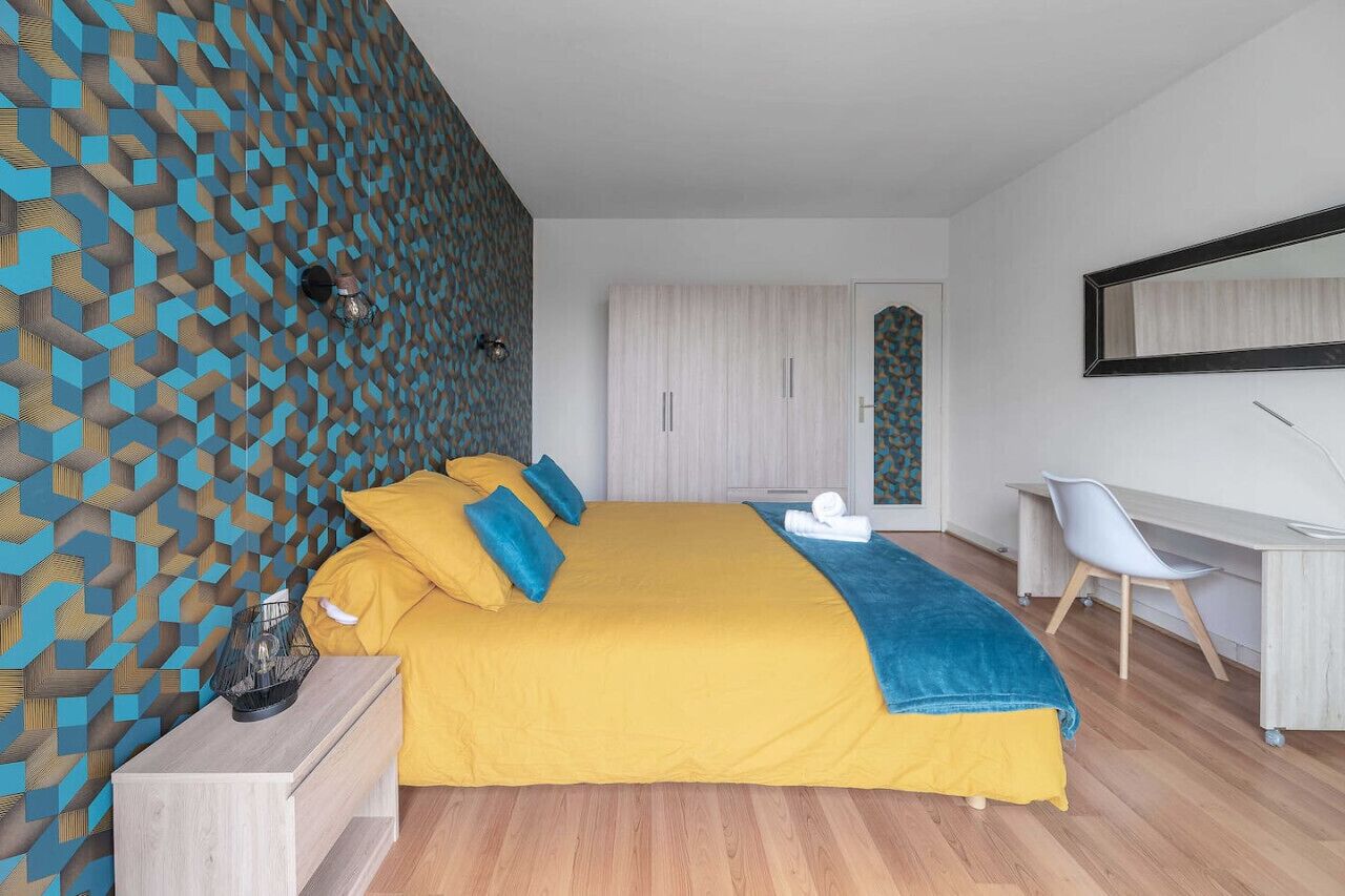 A modern bedroom with a geometric patterned wall, a large bed with yellow bedding, a desk with a chair, and a wardrobe.