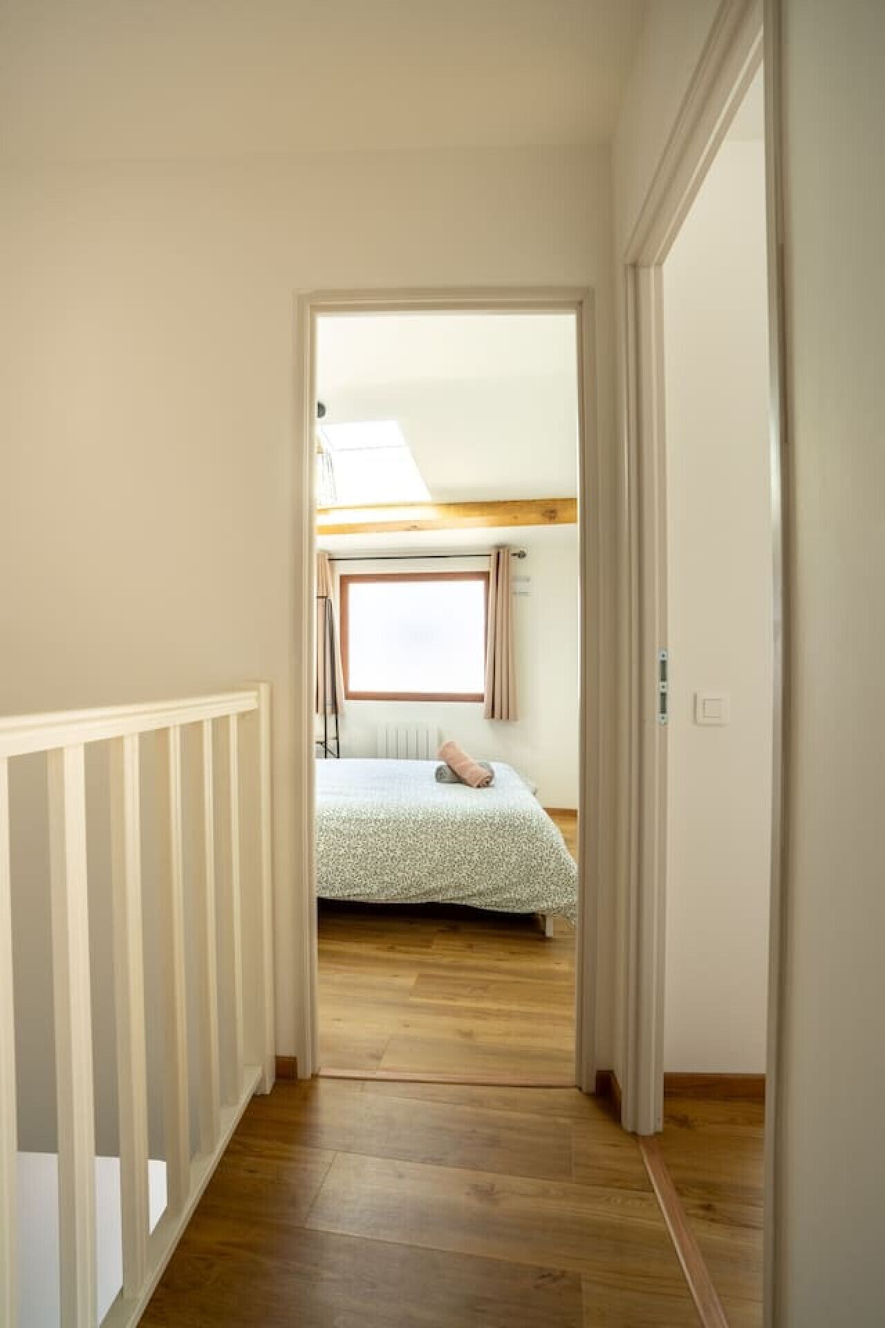Passadís estret que porta a un dormitori amb sostre inclinat, una finestra amb cortines i un llit amb mantell estampat.