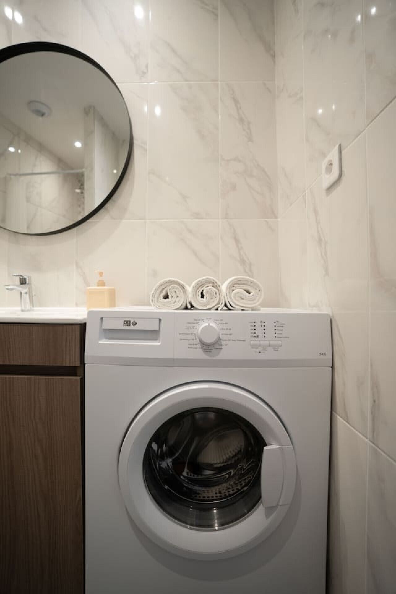 Een moderne wasruimte met een voorlader wasmachine, een houten kast, een spiegel en een betegelde muur van marmer.