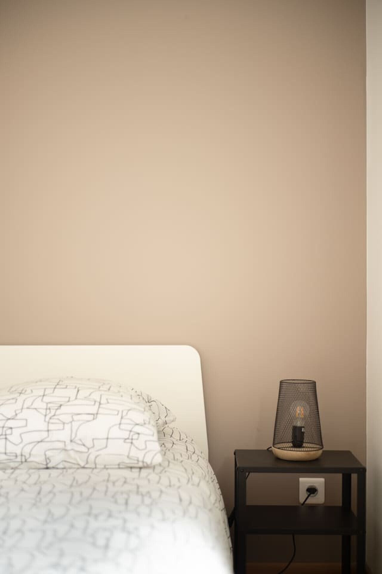 Ein ordentlich bezogenes Bett mit einer gemusterten weißen Tagesdecke und einem schlichten weißen Kopfkissen. Gegen eine schlichte Wand ist ein kleiner Nachttisch mit einer Netzleuchte und einer Steckdose gestellt.