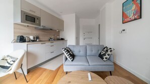 Ein modernes Wohnzimmer mit grauem Sofa, einem kleinen runden Tisch und einer Küchenzeile mit Mikrowelle und Kaffeemaschine.