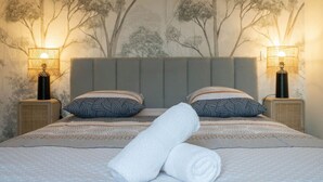 Ein ordentlich gemachtes Bett mit grauem Kopfteil, zwei gemusterten Kissen und einem aufgerollten weißen Handtuch. Auf jeder Seite des Bettes stehen je eine Nachttischlampe.