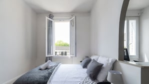 Ein Schlafzimmer mit Bett, Nachttisch, Lampe und einem Fenster mit geöffnetem Klappladen.