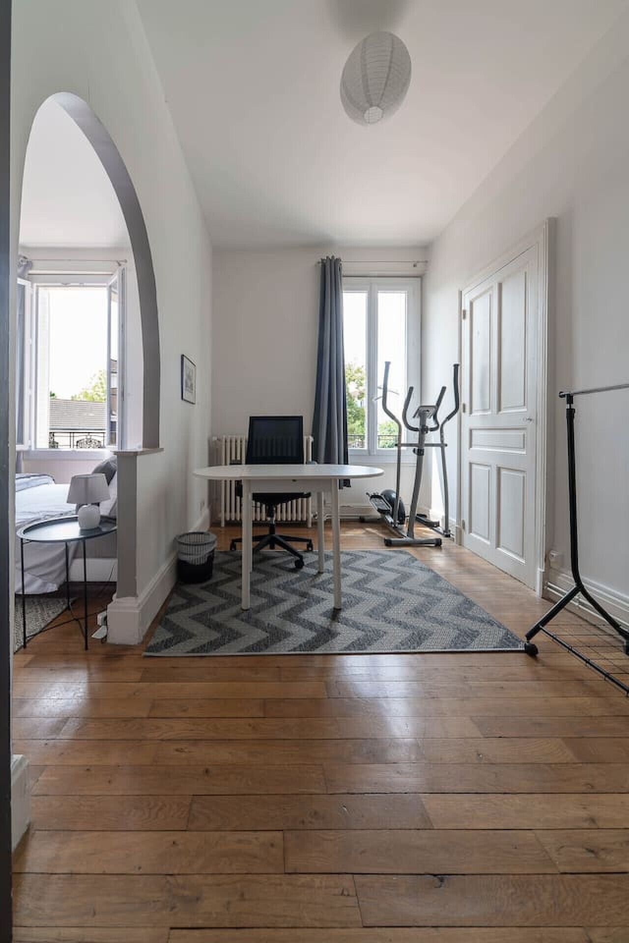 Une pièce avec un plancher en bois, un bureau blanc avec une chaise, un radiateur, un lampadaire et une fenêtre avec des rideaux.