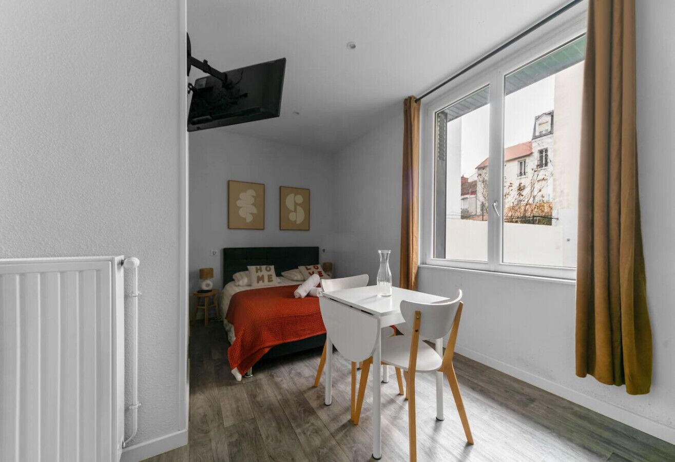 Una habitación con una cama, una mesa blanca con sillas, una ventana con cortinas y un radiador.