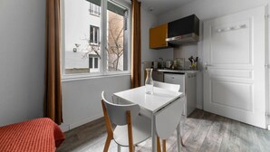A compact kitchen with a dining area, a window with curtains, and a view of a building outside. - Appartement design Centre Ville proche Thermes (Vichy)