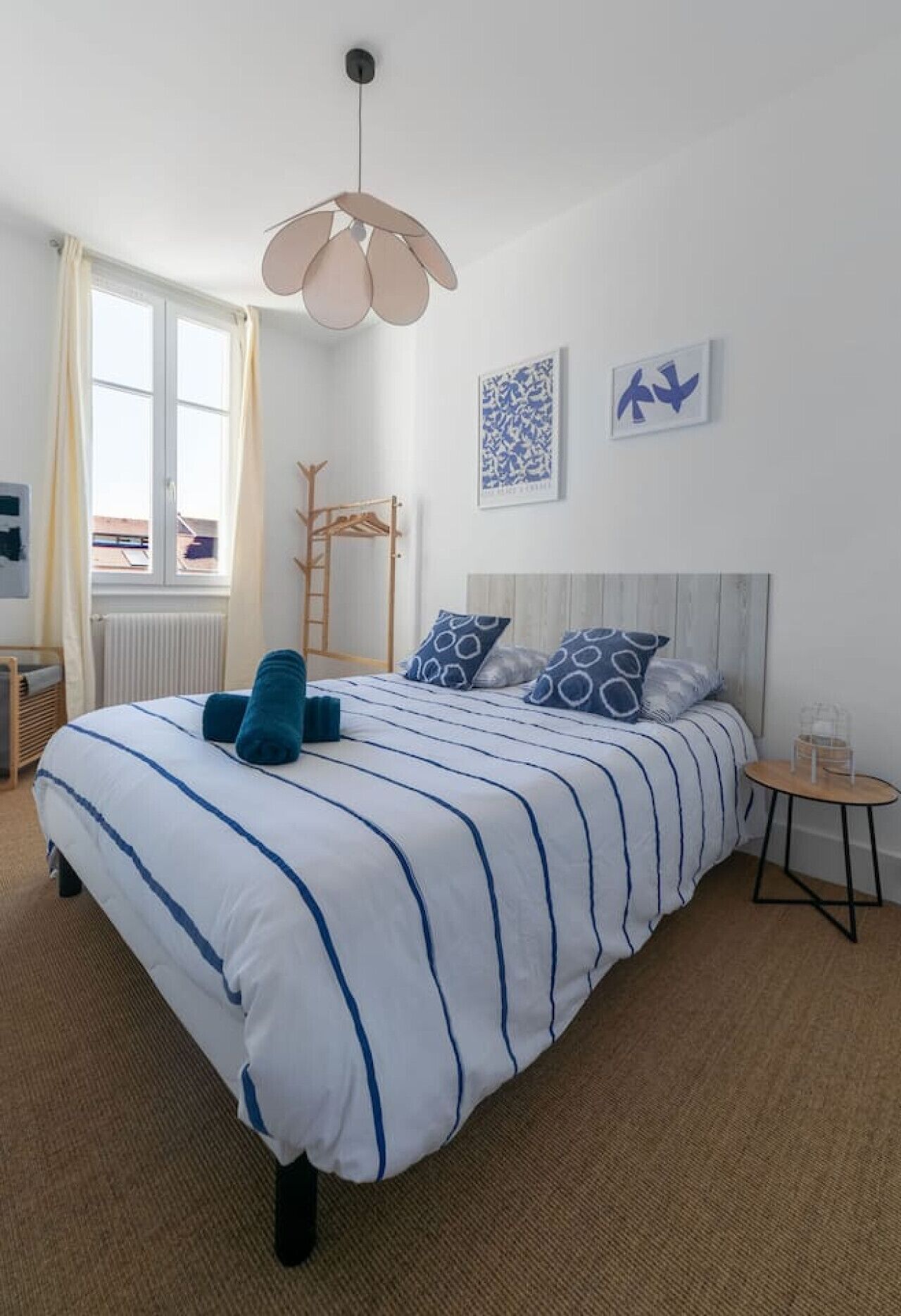 A neatly arranged bedroom with a large bed, a bedside table, a hanging light fixture, and a window with curtains.