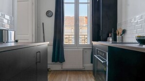 Cocina estrecha con armarios oscuros, una ventana con cortinas, un radiador y vistas a edificios.