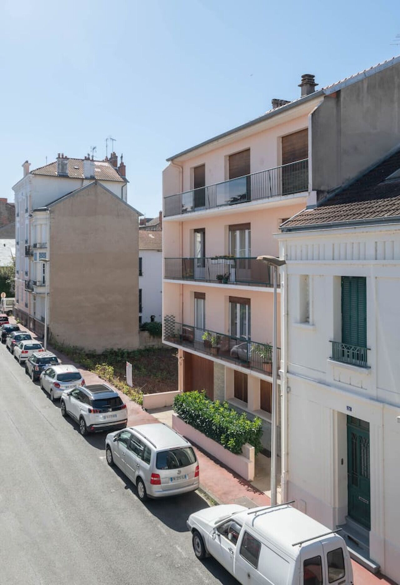 Una hilera de coches aparcados a lo largo de una calle con edificios de varios pisos, balcones y techo plano.
