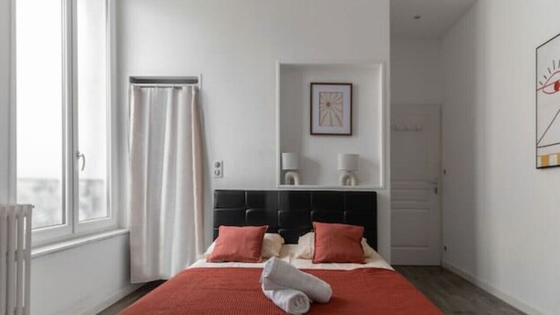 A modern bedroom with a large bed, a window with curtains, a headboard, a bedside table, and a framed picture on the wall.