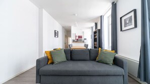 Una sala de estar moderna con un sofá gris, cojines amarillos y verdes, una mesa de comedor al fondo y un cuadro en la pared.