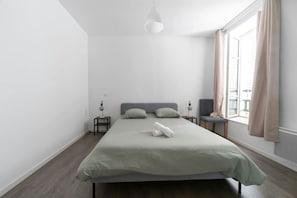 A minimalist bedroom with a large bed, two bedside tables, a chair, and a window with curtains. - Grand appartement design, 3 chambres, centre ville (Vichy)